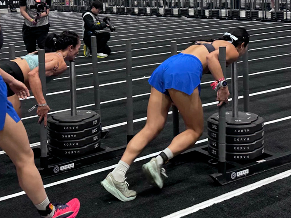 Athlete in blue shorts pushing a weighted sled with stacked plates during a workout, photographers in the background.