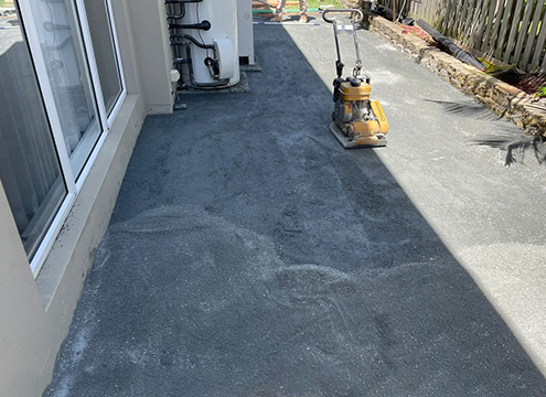 Yellow plate compactor on a freshly laid concrete patio beside the house, likely being resurfaced outside a doorway.