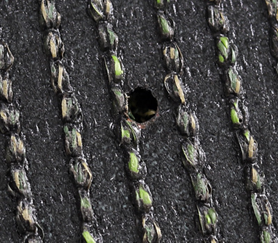 Close-up of a dark surface with parallel rows of small green-tinted stitches and a central hole (macro texture).