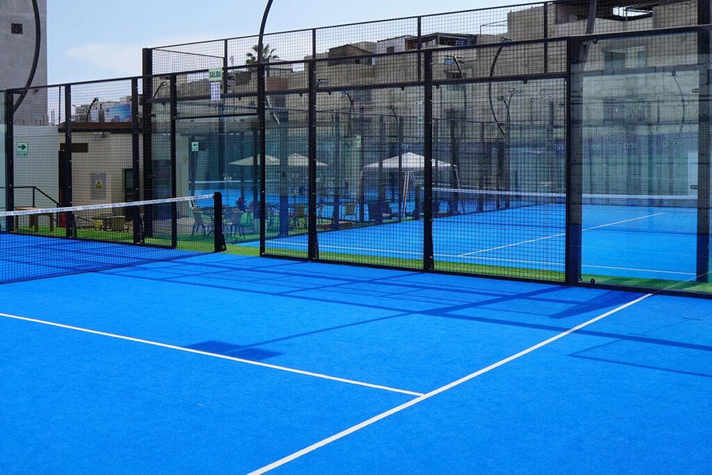 Blue outdoor padel/tennis court with a central net and black wire fencing, glass walls, and seating area in the background under a sunny sky