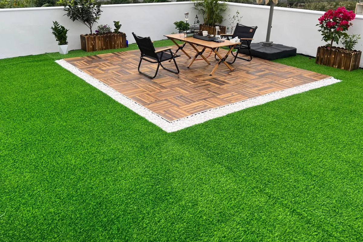 Outdoor patio area with a wooden tiled platform surrounded by white gravel and vibrant green lawn, table and chairs set.