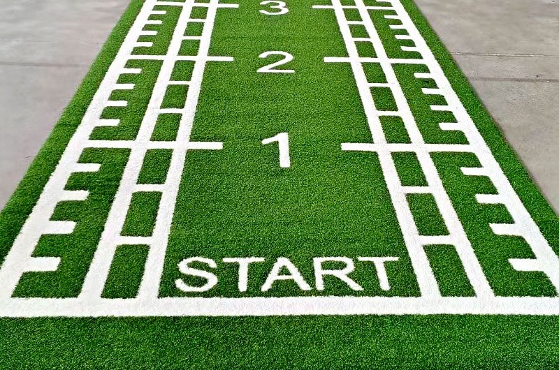Green artificial turf race starting line with bold white 'START' and lane numbers 1–3 visible from above.